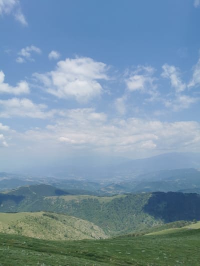 This media file depicts a vast, rolling mountain landscape under a bright blue sky dotted with white, puffy clouds. The foreground is a gently sloping grassy hill, lush with green grass and scattered small wildflowers. Further down the slope, the terrain becomes more rugged, with patches of more sparse vegetation and rocky outcrops.

The mid-ground and background are dominated by a series of verdant, tree-covered mountain ranges that recede into the distance. The forests appear dense and healthy, with varying shades of green indicating different types of foliage and perhaps the play of light and shadow. A deep shadow is cast across one of the prominent mountain slopes, suggesting the time of day is likely mid-morning or late afternoon.

In the far distance, a hazy, almost ethereal view of a valley or plain can be seen, dotted with what appear to be scattered settlements or buildings. The atmosphere is clear but with a slight haze that softens the distant details, creating a sense of depth and expanse.

The overall impression is one of natural beauty and tranquility. The scene is peaceful, evoking a feeling of serenity and the grandeur of nature. The weather appears to be pleasant and sunny, ideal for outdoor exploration. The location is identified as Brestovo, Bulgaria, suggesting a rural, mountainous setting. There is no visible text in the image. The image captures a moment of natural splendor, with the vast sky and expansive mountains dominating the frame.