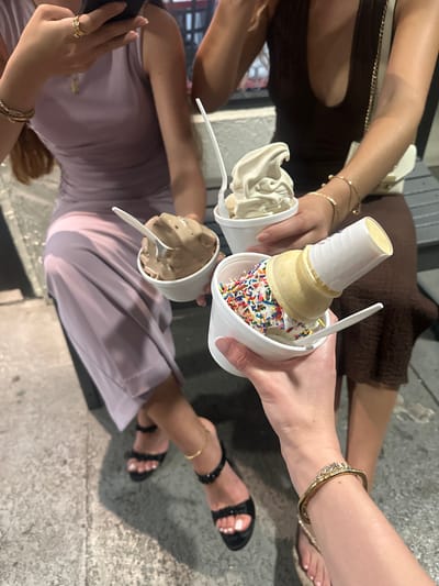 Three women are holding ice cream, sitting on a concrete surface, likely steps. One woman is holding a cup with an ice cream cone inside covered in sprinkles. One has chocolate ice cream, and one has vanilla ice cream. All the women are wearing skirts and jewelry on their arms, hands, and ankles. One woman has black sandals. The location is not discernible.