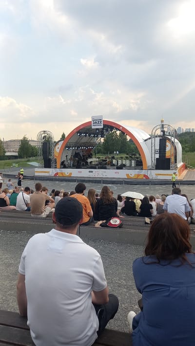 The image captures an outdoor jazz festival in the city of Путрышкаўскі сельскі Савет, Belarus. A stage with an orange archway is set up, featuring musical equipment and performers. Spectators are seated on tiered benches in the foreground, facing the stage. The sky is overcast with scattered clouds. The overall scene depicts a relaxed, community gathering focused on music.
