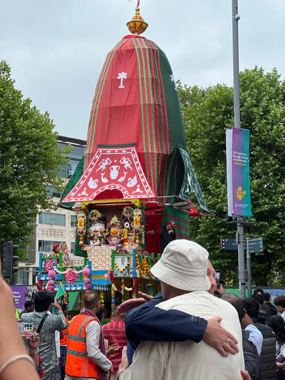 This is a medium shot, captured from a slightly low angle, of a religious procession in Belfast City District, United Kingdom. The central focus is a large, ornate chariot, draped in red and green fabric, with decorative elements on its upper structure. Several figures are visible on the chariot's platform, including statues adorned with garlands. A crowd of people surrounds the chariot, many with their phones raised, appearing to be spectators or participants. In the foreground, two men are embracing, one wearing a white bucket hat and the other with his back to the camera. The environment suggests an urban setting with trees and buildings visible in the background. A banner hangs from a lamppost on the right, displaying text in Irish and English, possibly indicating a festival or event. The overall mood appears festive and communal.