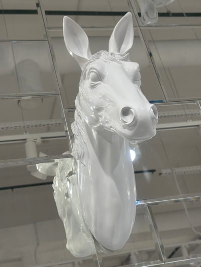A white horse-head sculpture, glossy and detailed, is displayed behind a glass and mirrored wall. The sculpture is positioned to the right and slightly above center. Its head features prominent ears, detailed facial features, and a smooth body. The background consists of multiple mirrored squares creating reflections. The mirrored structure has a grid pattern with visible hardware. The environment is predominantly white or light-colored, suggesting an indoor setting like a store or gallery. The location in the city of Kuleshovka, Russia, is not verifiable from the visual information provided.