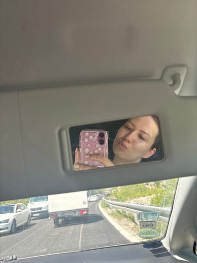 This vertical photograph captures a casual moment inside a car in Obzovica, Montenegro, during what appears to be bright daylight. The main subject is a young woman, visible in the vanity mirror embedded in the car's sun visor. She is taking a selfie, making a playful "kissing" face with puckered lips. Her hair is dark and pulled back, and her complexion is fair. She holds a pink smartphone with a clear case featuring white daisy patterns, which is also reflected in the mirror. Her fingernails are painted a light, neutral shade.

The car's interior, primarily the sun visor and headliner, is a muted gray. Through the windshield, a sunny outdoor scene unfolds, indicating clear weather. The car appears to be part of a queue or moving slowly in traffic on a road, possibly a highway, given the visible metal guardrail and other vehicles ahead, including a white truck and a silver car. Beyond the road and guardrail, lush green vegetation can be seen on an embankment.

Several official-looking stickers are affixed to the car's windshield. Prominently visible are "2021" and the logo for "DEKRA," a German vehicle inspection company, suggesting the car may have undergone inspection. Another sticker appears to display "12 2021," likely indicating a date. The bright sun casts distinct shadows, further emphasizing the daytime setting.