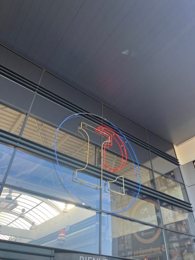 The image shows a large, modern building with a glass facade in Saint-Brice-Courcelles, France, a suburb of Reims. The building has a large, neon sign in the form of a circle with a letter "T" inside it. The neon is red, yellow, and blue. Behind the sign, a glass roof with a white steel structure and an array of large light fixtures can be seen. There are other large, modern signs with unintelligible text in the background.