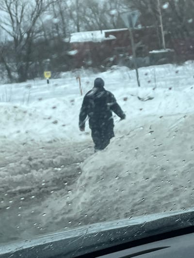The image depicts a winter scene in Rogachovo, Russia, observed from inside a vehicle through a wet windshield. A single individual, viewed from behind, is walking away from the camera through snow-covered terrain. The person is dressed in dark, possibly patterned, winter outerwear including a hooded jacket or coat and a dark beanie, and appears to be holding a long, thin object in their right hand, pointed towards the ground. The immediate foreground shows a significant accumulation of dirty, partially melted snow and slush on the right side, along with the lower edge of the vehicle's interior. The ground where the person is walking is covered in snow, with visible darker patches possibly indicating exposed earth or debris. In the mid-background, a small yellow sign is visible to the left, and a utility pole or similar structure stands to the right. The far background features leafless trees and a reddish-brown building with a snow-covered roof, partially obscured by the trees. The overall environment is overcast and appears cold, consistent with winter conditions.