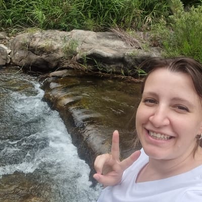 A woman, with brown hair and a white t-shirt, is taking a selfie outdoors. She is smiling and making a peace sign with her left hand. Behind her, a stream flows over rocks, with green foliage and more rocks visible in the background. The setting appears to be a natural environment, possibly a park or a hiking trail, consistent with the natural beauty of Mureungdowon-myeon, South Korea.