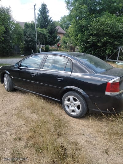 A black sedan is parked on a dry, grassy area next to a wooden fence and trees. The car is facing left, with its rear end partially visible. The background shows a house with a brown roof and more trees. The sky is overcast. A timestamp in the bottom left corner reads "2026.07.03 14:09". The scene appears to be in Novyi Myliatyn, Ukraine.