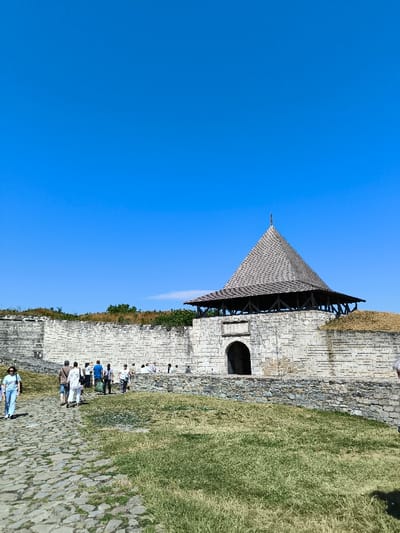 The image displays an outdoor scene at the Khotyn Fortress in Khotyn, Ukraine, under a clear, bright blue sky with a faint, wispy cloud visible near the horizon on the left. The lighting indicates a sunny daytime environment, with short shadows suggesting a high sun angle.

The central feature is a large, light-colored stone fortress wall extending horizontally across the mid-ground. Slightly to the right of center, a prominent stone gatehouse or tower rises from the main wall. This structure includes an arched entrance at its base, a weathered rectangular panel above the arch, and an exposed wooden superstructure that supports a tall, multi-tiered conical roof covered in light gray, possibly wooden, shingles. A small finial or spire crowns the apex of the roof. A lower, darker stone wall runs in front of the main wall, segmenting the foreground. Beyond the fortress walls, green foliage is visible along the distant horizon.

In the foreground, a cobbled stone pathway extends from the lower left, running alongside an area of sparse green and brown grass on the right.

Approximately 10 to 15 individuals, appearing as visitors, are present in the mid-ground. Most are observed walking along the stone path or standing near the base of the main fortress wall, primarily to the left of the gatehouse. One individual, dressed in a light blue top and light-colored pants, walks alone in the left foreground, moving towards the viewer. The individuals are dressed in casual attire. No specific interactions or detailed actions beyond walking and observation are clearly discernible among the groups.