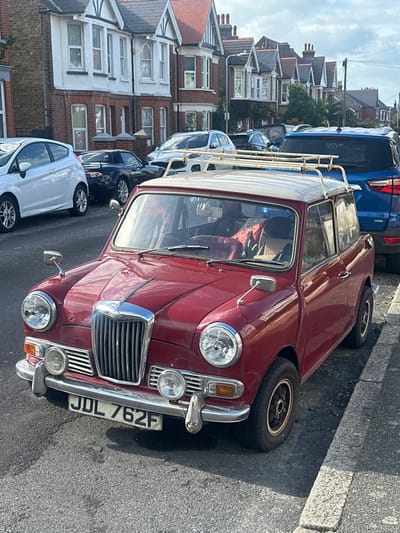 The image depicts a static street scene in Thanet, United Kingdom. In the mid-ground, a dark red vintage two-door car, license plate JDL 762F, is parked on the left side of an asphalt road, angled slightly towards the viewer. The vehicle features a cream-colored roof with a white roof rack, a chrome vertical grille, circular headlights, and two smaller auxiliary lights below the bumper. Its interior shows a red fabric item and two bottles on the front passenger seat. The road's edge is defined by a grey concrete curb on the right. In the background, a continuous row of multi-story terraced houses lines the street, characterized by brick ground levels, white upper facades, bay windows, and dark-tiled gable roofs. Other vehicles are parked along the street: a white hatchback is visible to the immediate left of the vintage car, followed by a dark blue sports car. A blue SUV is partially visible further down the street on the right. The sky is partly cloudy, and the scene is illuminated by daylight. No individuals, actions, or interactions are present.