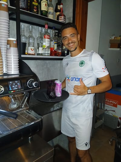 The image captures an indoor scene at a bar or cafe in Sotira, Cyprus. A young adult male, with short dark hair and a light beard, stands slightly right of center, smiling directly at the camera. He wears a white and grey patterned short-sleeved sports jersey with a green cloverleaf logo (Panathinaikos) on the chest, an "OPAP" sponsor logo below it, and a "Macron" logo on the right sleeve. He also wears white shorts with a "Macron" logo on the left leg and a gold-toned rectangular wristwatch on his left wrist. He holds a dark circular serving tray in his left hand, which contains two dark purple/brown glasses (one with ice) and two pink beverage cans. His right hand is extended, forming a "shaka" or "hang loose" gesture.

Behind the man, a black metal shelving unit against a grey textured wall displays an assortment of liquor bottles on two shelves. The top shelf includes bottles of Jack Daniel's Tennessee Whiskey, Passoa Passion Fruit Liqueur, Jameson Irish Whiskey, Lillet Blanc, and other liqueurs. The lower shelf features five Monin syrup bottles (including "Saveur Litchi" and "Triple Sec"), a Stolichnaya Vodka bottle, several clear glasses, an Aperol bottle, and another dark liquor bottle.

To the left, a tall stack of light-colored disposable coffee cups is visible. Below the shelves and to the left of the man is a black and chrome professional espresso machine, labeled "MADE IN ITALY," with a digital display showing temperatures (90.5°C, 90.3°C), the time (20:07), and the date (5/6/2026). A stainless steel counter surface is present below the machine and extending to the right, with an ice well visible behind the tray. A red and white "FILIPOU CREAM" branded box is partially visible in the lower right background. The area is well-lit by artificial indoor lighting.