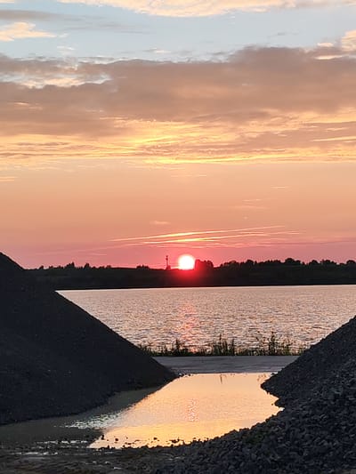 The image captures a serene sunset over a body of water, likely a lake or river, near Faliukovo, Russia. The sky is ablaze with warm hues of orange, pink, and yellow, transitioning into a soft blue at the top, with scattered, wispy clouds. A sliver of the sun is visible on the horizon, casting a vibrant pink glow. The distant shoreline is silhouetted against the sky, appearing as a dark band of trees and what might be a small structure with a tower. In the foreground, two large piles of dark aggregate material, resembling coal or gravel, frame the scene. A puddle of water between these piles reflects the colorful sky. The overall impression is one of natural beauty and tranquility, with industrial elements in the foreground hinting at a quarry or construction site. There are no people or animals visible in the image.
