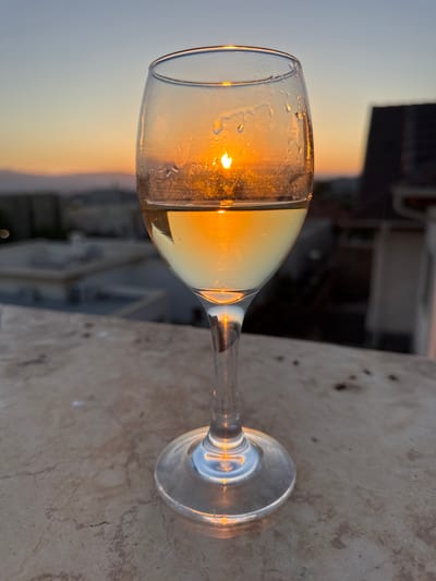 A close-up shot of a wine glass filled with a light-colored liquid, possibly white wine, is positioned on a textured, stone-like surface. The setting sun is visible through the glass, its golden rays creating a warm glow and reflecting off the liquid and the glass's condensation droplets. The background, slightly out of focus, reveals the silhouetted rooftops of buildings and distant hills, suggesting an urban or suburban environment. The sky displays a gradient of colors, transitioning from a soft blue at the top to warm oranges and yellows near the horizon, indicative of a sunset. The scene is likely located in Katzrin, Israel, given the architectural style and the ambient lighting.