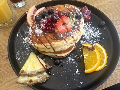 A close-up, high-angle shot depicts a stack of four pancakes served on a matte black circular plate. The pancakes are covered with a dark chocolate or hazelnut spread, liberally dusted with powdered sugar, and topped with whole blueberries, redcurrants, and a halved fresh strawberry. Arranged around the pancake stack on the plate are garnishes including a thin slice of grapefruit, two triangular slices of pineapple, two crescent-shaped orange slices, scattered whole blueberries, and a small cluster of red grapes. Additional dark syrup or chocolate drizzles are present on the plate surface. The plate rests on a light-colored wooden table. In the blurred background, a glass containing amber liquid (likely orange juice) is visible on the upper left, and a glass with a dark beverage is partially visible on the upper right. No individuals are present. The specific location in Weißenburg in Bayern, Germany, cannot be determined from the visual information within the image.
