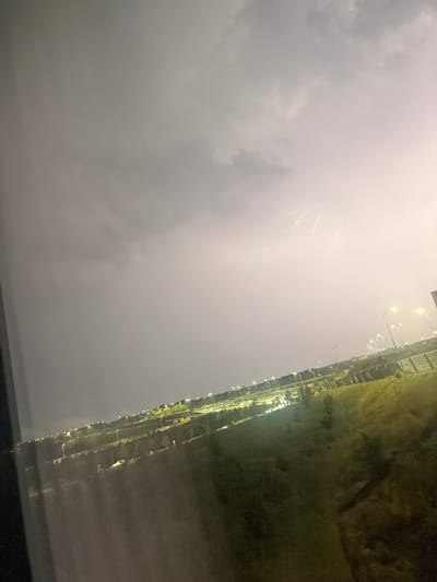 A nighttime thunderstorm illuminates the sky over the A9 highway and surrounding areas in Badhoevedorp, Netherlands. Jagged streaks of lightning flash against a backdrop of dark, ominous clouds. Streetlights line the highway, casting a warm glow on the road and the adjacent grassy slopes. In the foreground, a dimly lit window frame creates a slight obstruction, suggesting the photo was taken from indoors, possibly a residential building or hotel. The overall scene captures a dramatic natural event occurring within an urban landscape.