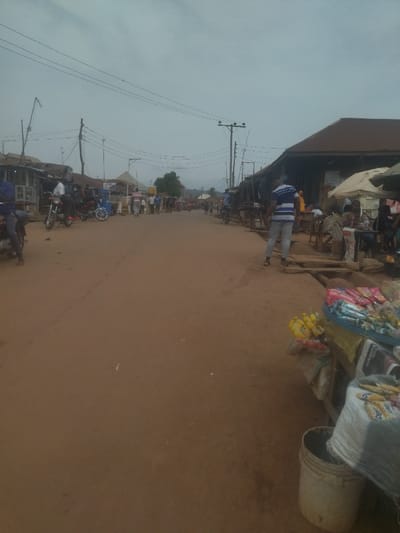 The image shows a dirt road in Junction, Nigeria, located at coordinates 8.958 latitude and 7.551 longitude. Several people are walking down the street in the distance, including a man in a blue and white striped shirt. There are also several motorcycles parked on the side of the road. In the foreground, there is a small stand selling candy and other goods. The stand is situated on the edge of the road next to a white bucket. The day is overcast, with a gray sky and a light mist in the air. Power lines run over the road and through the scene. The scene is a rural village setting in Nigeria with unpaved roads and small shops along the sides.