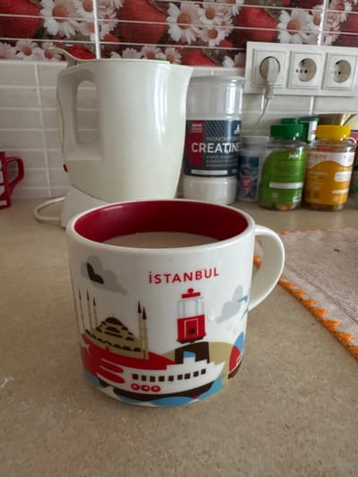This image, taken in a kitchen located in Ciorescu, Moldova, displays a white ceramic mug with a red interior rim, filled with a light brown liquid, positioned on a beige countertop. The mug features a multicolored graphic design depicting Istanbul landmarks, including a mosque-like structure, a ferry boat, and the red Tünel tram, with the word "İSTANBUL" printed in red above.
Behind the mug, from left to right, are several items: a white electric kettle with its power cord visible, a large clear plastic container labeled "CREATINE MONOHYDRATE" in black and red text, a smaller blue and white bottle, a green plastic bottle of supplements, and a yellow plastic bottle of supplements. To the right of the kettle and above the supplement containers, a white electrical outlet with three European-style sockets is visible on a white tiled backsplash. The backsplash features a repetitive pattern of red strawberries and white daisy-like flowers on a lighter background. A small section of a beige fabric cloth with an orange ruffled edge is visible on the countertop to the far right. The scene is brightly lit. No individuals are present.