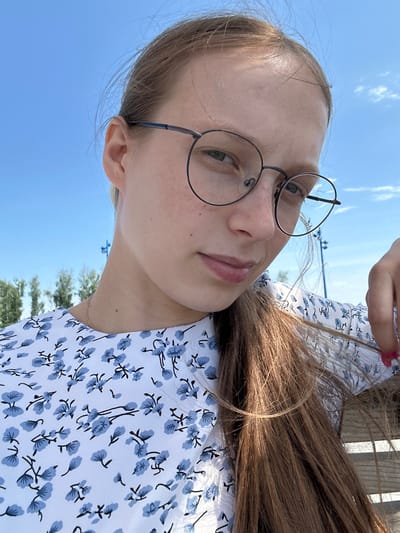 A young woman with long, brown hair is taking a selfie outdoors. She is wearing round, dark-rimmed glasses and a white dress with a blue floral pattern. Her hair is pulled back into a ponytail and falls over her shoulder. She is looking at the camera with a slight smile. The background shows a bright blue sky with a few clouds, and some green trees and structures are visible in the distance. The location appears to be a park or open area, possibly on the outskirts of the city.