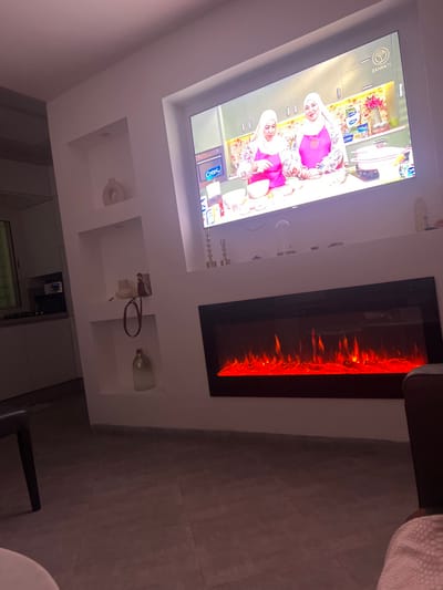 A contemporary living room features a recessed television showing a cooking show with two women wearing hijabs. An electric fireplace with simulated flames is mounted next to the TV. Below the TV and fireplace are built-in shelves displaying decorative items, including candles, vases, and abstract art. The room is modern with tiled flooring and muted lighting. A partial view of furniture is visible in the foreground.