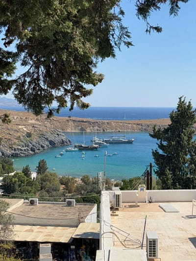 A scenic, sun-drenched view of Lindos, Greece, showcases a picturesque bay filled with various boats, including yachts and smaller vessels. The water is a vibrant turquoise, inviting and clear, with the rocky coastline of the ancient city serving as a dramatic backdrop. Lush green trees and foliage dot the landscape, contrasting with the warm, earthy tones of the arid hills. In the foreground, a rooftop of a building is visible, featuring air conditioning units and outdoor furniture. A glimpse of what appears to be shops or stalls is also present at the bottom of the frame. The sky is a clear, brilliant blue, indicating fair weather.