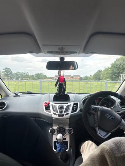 The image provides an interior view from a stationary Ford Fiesta, looking forward through the windshield at a green recreational field in Boston, United Kingdom.

**Car Interior:** The dashboard is dark grey with silver accents around the central console and air vents. On the driver's side, a black, textured steering wheel featuring a carbon fiber pattern wrap and a blue stripe displays a Ford emblem. The instrument cluster indicates "20.6 mi" on its digital display. The legs of an individual, wearing light-colored trousers and partially obscured by a cream-colored, textured blanket, are visible in the driver's footwell. On the passenger's side, an individual's legs clad in black ribbed trousers are present. The central console contains a radio/CD player unit with various buttons (e.g., RADIO, CD, AUX, PHONE, MENU, OK) and three circular climate control knobs. A manual gear stick has a blue cloth item tucked at its base. A red, bone-shaped ornament is attached to a passenger-side air vent. A black phone mount is affixed to the dashboard near the windshield. The rearview mirror reflects the top of a person's head. Several items hang from the rearview mirror, including a red fluffy pom-pom, multiple keychains, and a small black boxing glove. The car's grey headliner and raised sun visors are visible overhead.

**Exterior Environment:** Outside the vehicle, a grey metal fence with vertical bars separates the car from a large, bright green grassy field. Towards the left of the field, distant playground equipment, including swings and slides, is visible. In the background, a cluster of red-brick residential buildings, including a more prominent, multi-windowed structure, stands beyond the field. Abundant green trees with full foliage line the distant horizon and are interspersed among the buildings. A white goalpost is partially discernible on the far right. The sky is uniformly overcast with white and grey clouds.
