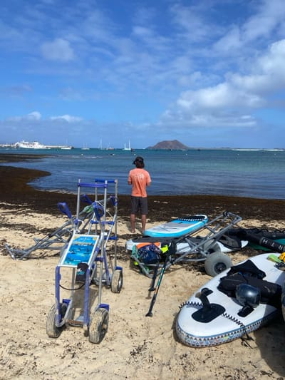 The image shows a sandy beach in Corralejo, Spain (latitude: 28.734, longitude: -13.867). One person is standing on the beach facing away from the camera, while a second person is partially obscured by the cart. The person standing is wearing a pink shirt and blue shorts. The person on the cart is wearing a black shirt and black shorts. There are three carts in the image, and the cart furthest from the camera has a blue and silver frame with an advertisement on it. The other two carts are similar in color and design. The beach is covered in seaweed and a few small boats can be seen in the distance, as well as a large mountain in the background. The sky is blue with white clouds.