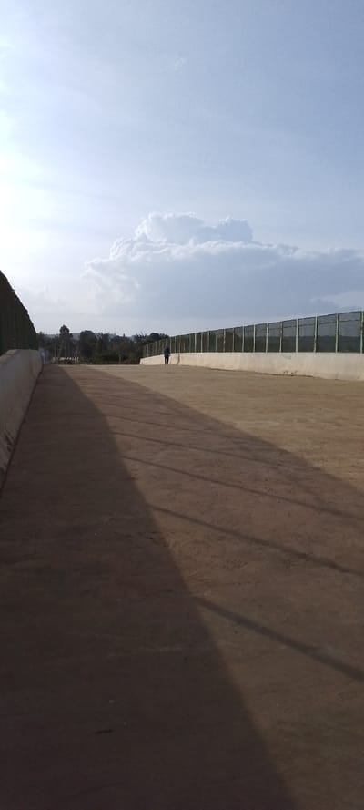 The image captures an expansive outdoor scene, likely a wide pathway, road, or open area, under a bright sky with scattered clouds. The ground is a light brown or tan color, possibly a packed dirt or gravel surface, with long, distinct shadows stretching across it from left to right. These shadows are cast by elements outside the frame and by the green barriers running along the sides, indicating either early morning or late afternoon light.

On both the left and right sides of the pathway, there are low, light-colored concrete or stone walls. Perched atop these walls are green fences or barriers that appear to be made of a mesh material or have semi-transparent panels. These barriers extend into the distance, suggesting a long, linear structure.

In the mid-ground, a single individual is visible, walking away from the viewer towards the left side of the frame. They are small in the distance, dressed in what appears to be dark clothing.

Beyond the barriers on the left, distant green foliage, possibly trees or hills, can be seen under the bright horizon. The right side shows more of the fence structure extending into the background. The sky above is a clear, light blue, filled with soft, white cumulus clouds, suggesting fair weather. The overall impression is one of open space and quiet, with the long shadows adding a sense of depth and time. There is no visible text in the image.