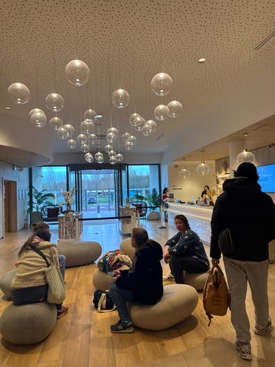 An indoor scene in Marcq-en-Barœul, France, depicts a modern lobby or waiting area. The foreground features a light-colored wooden floor and several people. On the far right, a person in a black hooded jacket, grey sweatpants, and white sneakers walks away from the camera, carrying a tan backpack in their right hand and a black fanny pack at their waist. In the mid-ground, a woman with dark hair, wearing a patterned dark blue top, sits on a light grey, bean-shaped ottoman, looking left. To her left, another woman with long dark hair, in a beige textured top and dark pants, sits with her back mostly to the camera on a similar ottoman, with a light green shoulder bag next to her. Another person is partially visible behind her. A dark blue tote bag rests on the floor.
The ceiling is white and subtly textured, from which numerous clear, spherical glass pendant lights of varying sizes hang at different heights. In the background, a large glass revolving door provides a view to the outside, showing an overcast sky, faint green foliage, and a distant road with a white car. To the right of the revolving door, a light-colored reception desk is staffed by two individuals. One person in a red top and white face mask stands behind the counter, facing left. Another person with dark hair, wearing a dark top, is visible further right behind the desk, with a baguette on the counter next to them. A sign above the reception desk reads "SANITAIRES." A large digital display screen with white text on a blue background is mounted on the wall above the desk. Several large potted plants, including palm-like varieties, are strategically placed around the area. A small wooden display stand with white flowers is located near the revolving door.