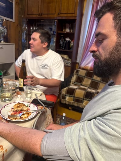 In a dimly lit interior, possibly a residential dining area, two individuals are seated at a table laden with food and drinks. The man in the foreground, with a full beard, wears a light blue t-shirt and has a blood pressure cuff wrapped around his upper left arm, suggesting a medical measurement is being taken. His right arm rests on the table, and his gaze is directed downwards.
To his left, another man in a white t-shirt, with dark hair, holds a brown bottle of beverage. He is looking away from the camera, his expression unreadable. A partially eaten meal sits on a plate in front of him, accompanied by glasses and a fork. The background reveals wooden cabinets, shelves with bottles and decorative items, and a patterned throw blanket draped over furniture. The presence of a blood pressure cuff implies a scenario where health monitoring is taking place within a casual social gathering. The overall atmosphere suggests a domestic setting, likely in the city of Ust-Mana, Russia, given the context provided.