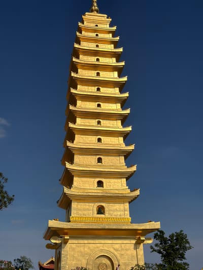 A vertical eye-level shot features the towering golden pagoda of Bái Đính Temple in Diên Lạc, Vietnam, set against a backdrop of deep blue sky. The pagoda's tiered structure, constructed of what appears to be gilded material, ascends from the lower right corner to the upper left, tapering towards its pinnacle. The intricate design of each tier is visible, including the recessed openings and architectural embellishments. To the right, sections of the temple's structure are visible, revealing round and detailed architectural features. Green foliage of trees frames the bottom right corner of the image. The photograph's composition emphasizes the pagoda's verticality and grandeur against the clear sky.