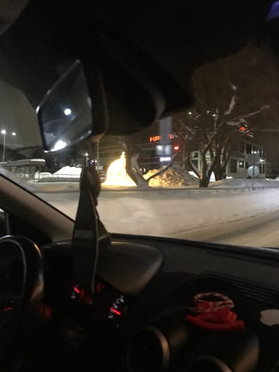 The image presents a nocturnal winter scene observed from the interior of a vehicle in Kremyonki, Russia.
Inside the vehicle, a dark, rectangular air freshener, potentially branded "AVON," hangs from the stem of a rearview mirror, which reflects a circular light source. A segment of a dark steering wheel is visible on the lower left. The dashboard displays red-illuminated gauges and, on its right side, a small red coiled object.
The exterior landscape is entirely covered in snow. The road surface appears partially cleared, contrasting with deep snow accumulation on the verges and sidewalks, forming several mounds. In the mid-ground, a large, spherical or dome-shaped decorative light installation emits a bright, warm yellow glow. Behind this, bare deciduous trees are heavily laden with snow. A multi-story building features red illuminated Cyrillic signage, prominently displaying "КЕ," likely part of "КЕНТАВР" (KENTAVR), a common name for commercial establishments. Another building with illuminated windows is visible further right. A snow-covered bus stop shelter is present on the far left, and a circular road sign is partially visible on the right side of the road. The scene is illuminated by ambient streetlights. No individuals are discernible.