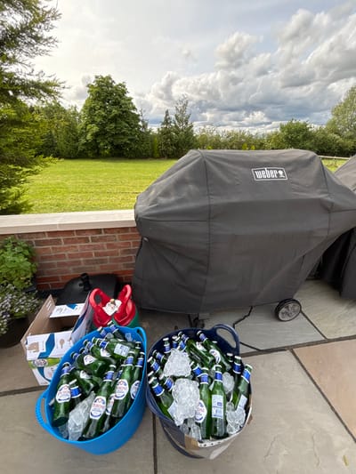 The image depicts a backyard scene in Newton upon Derwent, United Kingdom, with a brick wall in the foreground. Two blue plastic buckets filled with glass bottles of beer and ice sit on the patio. One bucket is labeled "Peroni." A black grill cover sits on top of a grill, with a "Weber" logo visible. A red and green propane tank sits beside the buckets. The sky is cloudy, with some areas of blue visible. The image suggests a social gathering or outdoor party.