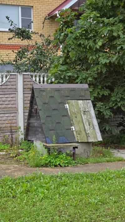 A well with a wooden roof sits in a grassy area next to a concrete fence. The well is constructed of concrete with a pitched wooden roof covered in green asphalt shingles and a weathered wooden door. A rustic wooden bench is positioned in front of the well. In the background, a yellow brick building with a red roof is visible, along with a large, leafy green tree. The scene appears to be in an outdoor residential or garden area.