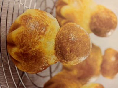 The image shows a close-up of several golden-brown baked goods, likely brioche or rolls, arranged on a wire rack. The buns have a soft, textured surface with some areas of lighter and darker browning. In the foreground, two buns are prominently featured, with one slightly larger than the other and attached. The background is blurred, showing more of the baked goods and the grid pattern of the wire rack. The lighting is warm and casts soft shadows. The image composition is a high-angle, close-up shot. The scene is located in a kitchen or bakery setting in Rumaithiya, Kuwait.