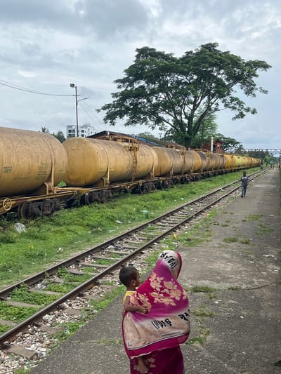 A low-angle, rotated shot captures a railway line in Sreemangal Upazila, Bangladesh. The foreground is dominated by a train consisting of numerous yellow, cylindrical tank cars, their surfaces appearing dusty. To the right of the train, a concrete platform runs alongside the tracks, scattered with debris. A person, whose body is partially visible, is dressed in a colorful pink and floral patterned garment, with the garment draped over their head. This individual is standing near the platform edge, close to the train.

Further down the tracks, a lone individual in dark clothing walks away from the camera. The train cars are lined up consecutively, stretching into the distance. To the left of the train, lush green grass grows alongside the tracks, and a large tree with dense foliage stands prominently. In the background, a few buildings are visible, along with a utility pole supporting lights and wires. The sky is overcast with grey clouds. The overall impression is a utilitarian scene at a railway station, likely during the daytime, given the lighting conditions.
