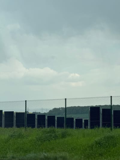 This image captures a landscape in Bad Rappenau, Germany, featuring a solar farm under an overcast sky. The upper half of the frame is dominated by a light grey to off-white cloudy sky, indicating diffused lighting conditions.
A wire mesh fence, with dark vertical support posts, runs horizontally across the mid-ground, acting as a boundary. Behind this fence, numerous dark, rectangular solar panels are arranged in rows, signifying a solar power generation facility. In the far background, beyond the solar panels, a line of dark green hills or dense tree lines forms the distant horizon. Faintly visible in the background, among the hills, are structures resembling utility poles or power lines.
To the right side of the frame, occupying the foreground, is a dense patch of lush, tall green grass. No specific activity or event is taking place, and no people are visible. The time of day appears to be either morning or afternoon on a cloudy day, given the soft, uniform light. There is no discernible text within the image. The scene presents a blend of modern renewable energy infrastructure with a natural, tranquil environment.