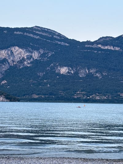 The image shows a wide shot of a serene lake with a mountainous backdrop in Conjux, France. The foreground features a pebble beach, with the calm, rippling surface of the lake occupying the middle ground. Two small objects are visible on the water: a red, flat object which appears to be a person or a watercraft, and a yellow buoy. In the background, a densely forested mountain range stretches across the horizon, with a small village nestled at its base. The sky is a clear, pale blue, suggesting fair weather.