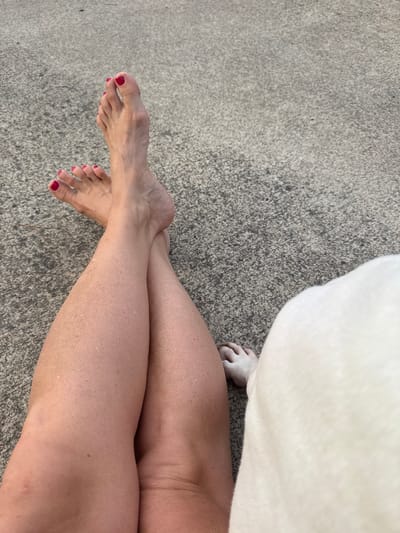 The image shows a person's legs and feet crossed over each other, resting on a textured, gray surface, possibly pavement or concrete. The toes of both feet are painted with a bright red nail polish. The skin on the legs appears to have some small bumps or texture. In the lower right corner of the image, a white furry object, possibly a blanket or a pet, partially covers another foot, which has white fur and a small pink toe. The setting appears to be outdoors. There is no specific indication of the city of Cartersville, United States, in the image itself.