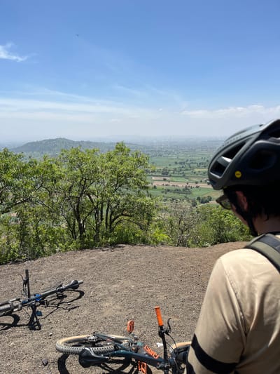 This image depicts a scenic overlook from a hillside, likely in the vicinity of San Miguel Papaxtla, Mexico. In the foreground, two mountain bikes are resting on a gravelly surface. One bike is upright and appears to be secured with a kickstand, while the other is lying on its side. A person wearing a beige shirt and a dark helmet is visible from the shoulders up, facing away from the camera and looking out at the expansive landscape. The environment features lush green trees and vegetation in the mid-ground, with a hazy view of a city or town spread across the valley in the background. The sky is bright blue with scattered clouds, and a tiny, unidentified flying object can be seen in the upper portion of the frame. The overall scene suggests a pause during a bicycle ride, taking in the panoramic view of the rural and urban expanse.