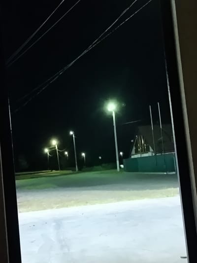 The image depicts an outdoor scene at night, possibly on the outskirts of a settlement. In the foreground, a paved surface is visible, reflecting the ambient light. Beyond this, a grassy area leads to a road or pathway lined with streetlights. Several of these streetlights are illuminated, casting a greenish-white glow on the surroundings. In the background, a building with a distinctive A-frame roof and green lower section is visible, with three vertical poles in front of it. Power lines are strung across the dark sky. The overall impression is one of a quiet, sparsely populated area at night.