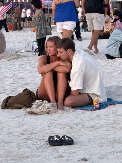The image captures a sandy beach scene at Cenang Beach, Malaysia, during what appears to be an evening gathering. In the foreground, a light-skinned female and male are seated directly on the sand. The female, positioned on the left, wears a dark, possibly black, swimsuit top. Her blonde hair is pulled back, and her arms are crossed over her chest; she gazes to her left with a neutral expression. The male, seated to her right, wears a light-colored, short-sleeved collared shirt and patterned shorts, and is turned towards the female. His left arm rests on his bent left leg, and he looks at the female. He sits on a blue towel with a red edge. A yellow SKOL beer can rests on the sand next to his right leg. A pair of white athletic shoes, visibly dusty, and a dark brown woven bag are on the sand near the female's feet. In the immediate foreground, a pair of black sliders with white "CK" (Calvin Klein) branding are placed on the sand. The background is populated by numerous out-of-focus individuals, including one holding a Malaysian flag on the far left, a person wearing a blue halter top and white shorts, a person with a black backpack, and a person in a hijab. Distant string lights and market stall structures are partially visible in the upper background.