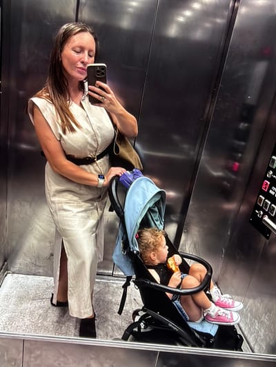 In a metallic elevator with reflective walls, a woman with long brown hair, wearing a beige dress and black belt, stands and takes a selfie with her phone. She is holding the handle of a stroller in which a young child, with curly hair, sits eating. The child wears a black top, denim shorts, white socks, and pink sneakers. The elevator has a control panel with illuminated buttons.