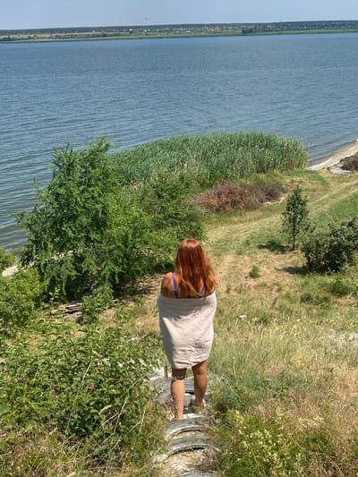 A woman with long, reddish-brown hair stands with her back to the camera, looking out over a body of water. She is wearing a light-colored, towel-like garment draped over her shoulders and a blue bikini top. The scene is a grassy, slightly overgrown embankment overlooking a wide, calm river or lake. Tall reeds grow along the water's edge. In the distance, across the water, a town or village with scattered buildings is visible along a treeline. The sky is clear and blue, suggesting a sunny day. The lighting indicates it is likely daytime, possibly late morning or afternoon, with the sun casting shadows on the grassy slope. The overall atmosphere appears peaceful and relaxed. The woman is positioned on a rough, circular object that resembles a concrete pipe or culvert, which is partially embedded in the ground and covered with vegetation. The setting is natural and somewhat wild, with a mix of green grass, dry brush, and trees.