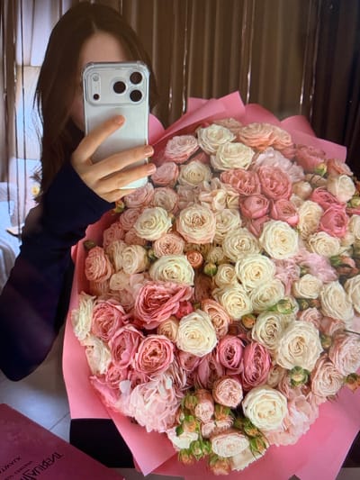 A woman, partially visible and holding a smartphone, is taking a picture of a large bouquet of pink and white roses. The bouquet is wrapped in pink paper. The background consists of a vertical brown blind. A pink book with Cyrillic text is partially visible in the bottom left corner of the image. The location is specified as Konstantinivka, Kyrgyzstan.