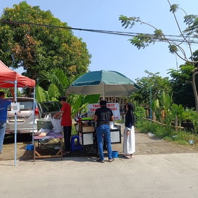 In Kebondalem, Indonesia, an outdoor food stall operates under a large green umbrella. The stall is positioned next to a silver car and a red canopy. Several individuals are present, including two men in casual attire, one in a blue t-shirt and the other in a red t-shirt, both appearing to be interacting with the stall or its products. A woman wearing a hijab and a long white skirt stands nearby. The stall itself is a cart with a display of food items and a sign that reads "TAHU KUROT TAHU GORENG UDANG" (Fried Tofu, Fried Shrimp Tofu). Bags of what appear to be snacks or ingredients are laid out on a table in front of the car. The environment is semi-urban with trees and vegetation visible in the background, under a clear blue sky. Power lines stretch across the scene.