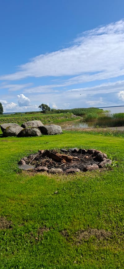 An outdoor scene in Valma, Estonia, features a circular fire pit in the foreground, filled with ash and charred logs. The pit is surrounded by large stones. Behind the fire pit, a meticulously mowed green lawn extends towards a body of water. Several large, moss-covered boulders are scattered across the lawn. In the background, tall reeds grow along the water's edge, with a distant shoreline visible. The sky is a vibrant blue, dotted with fluffy white clouds. A few birds are visible in the sky, and a single tree stands on a grassy knoll in the mid-ground.