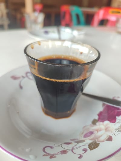 In Cotglumpang, Indonesia, a close-up shot focuses on a glass of dark coffee placed on a floral-patterned saucer. The coffee, dark and rich, fills the glass, with a thin layer of crema visible at the top. A spoon rests on the saucer to the right of the glass, its metallic sheen catching the light. The saucer itself is white with intricate floral designs in shades of pink and purple, with a subtle rim. The background is softly blurred, showing hints of furniture and other objects on a table, suggesting an indoor setting, possibly a cafe or a dining area. The lighting appears to be natural, casting a gentle glow on the scene.