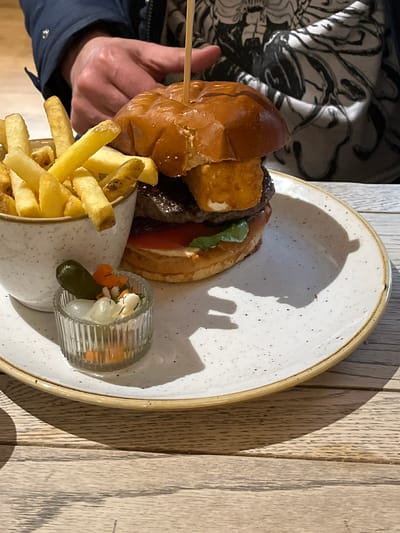 A close-up, eye-level shot captures a meal served on a light-colored wooden table within the Borough of Runnymede, United Kingdom. The central subject is a large burger, held by a wooden skewer, placed on a white, speckled ceramic plate with a light brown rim. The burger components include a glossy brioche bun, a beef patty, a square fried cheese piece, a red tomato slice, and green leafy vegetables. To the left of the burger, a white, speckled ceramic bowl holds golden-brown french fries. Adjacent to the fries bowl, a small, clear, fluted glass dish contains a single gherkin, several pickled white onions, and orange pickled carrot pieces. In the blurred background, a person's right hand is visible, positioned above the burger, wearing a dark blue jacket sleeve and a light-colored garment with an abstract black pattern. Direct lighting from the left casts distinct shadows of the food items and the hand onto the plate and table surface.