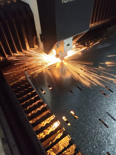 This is a close-up, high-angle shot of a laser cutting machine in operation, likely within an industrial setting in Dangir, Uzbekistan. The primary action is the laser beam intensely cutting through a metal sheet, indicated by the bright, energetic shower of sparks erupting from the point of contact. The sparks are predominantly orange and yellow, radiating outwards in a starburst pattern.
The machine itself is dark gray and black, with a visible white component holding the laser nozzle. Below the cutting area is a textured, dark gray metal surface with numerous elongated cutouts, suggesting a patterned sheet of metal is being processed. To the left, a ribbed, dark structure with a metallic sheen is visible, likely part of the machine's support or containment system. This structure also appears to be catching some of the sparks. The overall lighting is dominated by the bright glow of the sparks, creating strong contrast with the darker elements of the machine and the metal sheet. There are no people visible in the frame. The scene is focused on the industrial process of metal fabrication.