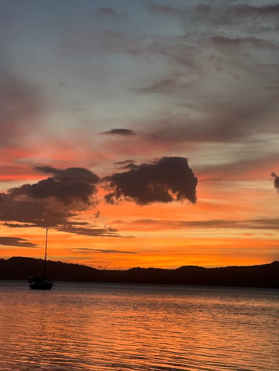 A sailboat is anchored on the calm waters of Lake Nicaragua, with the silhouettes of the Mombacho volcano and surrounding hills forming the horizon in the background. The sky above is ablaze with the vibrant colors of a sunset, transitioning from soft grays and pinks at the top to fiery oranges and yellows closer to the horizon. Wispy clouds are scattered across the sky, catching the last rays of the sun. The water reflects the dramatic colors of the sky, creating a shimmering, orange-hued surface. The scene is peaceful and serene, capturing the beauty of a sunset over a natural landscape in Altagracia, Nicaragua.