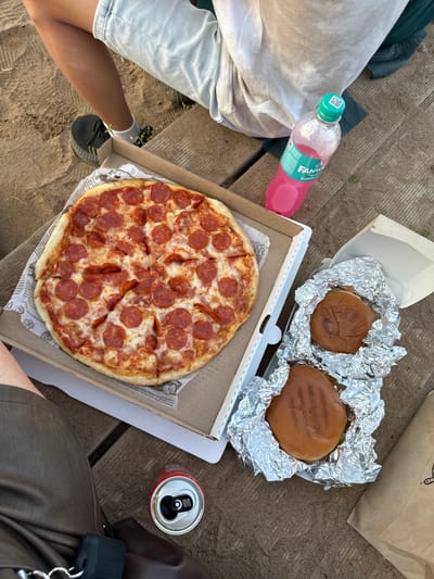 In Kostovaty, Russia, a casual outdoor meal is laid out on a wooden picnic table. A large pepperoni pizza, still in its cardboard box, sits open. To its right, two burgers are individually wrapped in aluminum foil. A pink bottle of Fanta Bubble Gum soda stands nearby, and a discarded red soda can rests on the table. The scene is partially framed by the legs of individuals wearing light-colored shorts and dark pants, indicating a relaxed setting. The ground appears to be sandy or earthy, consistent with an outdoor recreational area.