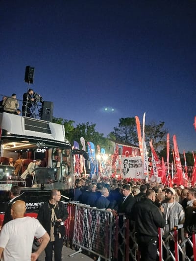 The image depicts a crowd of people gathered in Silivri, Türkiye, at the intersection of İstasyon Cad. and Belediye Meydanı. The scene is illuminated by artificial light. A white bus with the letters "CHP" emblazoned on its side is parked in the center of the frame, flanked by a crowd of people. The bus is a focal point of the scene, and it appears as if people are entering and exiting the vehicle. The majority of the crowd is dressed in casual clothing, while some wear more formal attire. There are numerous Turkish flags in the background, along with red and white banners. The flags and banners are displaying the image of Mustafa Kemal Atatürk, the founder and first president of the Republic of Turkey. Some individuals hold signs that read "Cumhuriyet Halk Partisi," which is the Turkish political party represented by CHP. The scene has a sense of excitement and anticipation, as people are seemingly gathered for an event or rally. The scene is captured in a low-light environment, likely during nighttime hours. The clear night sky with visible stars suggests a calm and quiet atmosphere. The image conveys a sense of political unity and shared purpose.